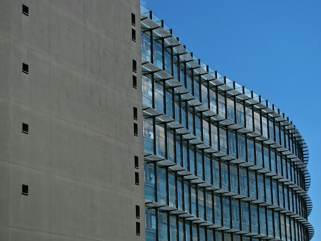 Modern curved glass building with sunshades next to a plain concrete wall with small square windows under a clear blue sky.