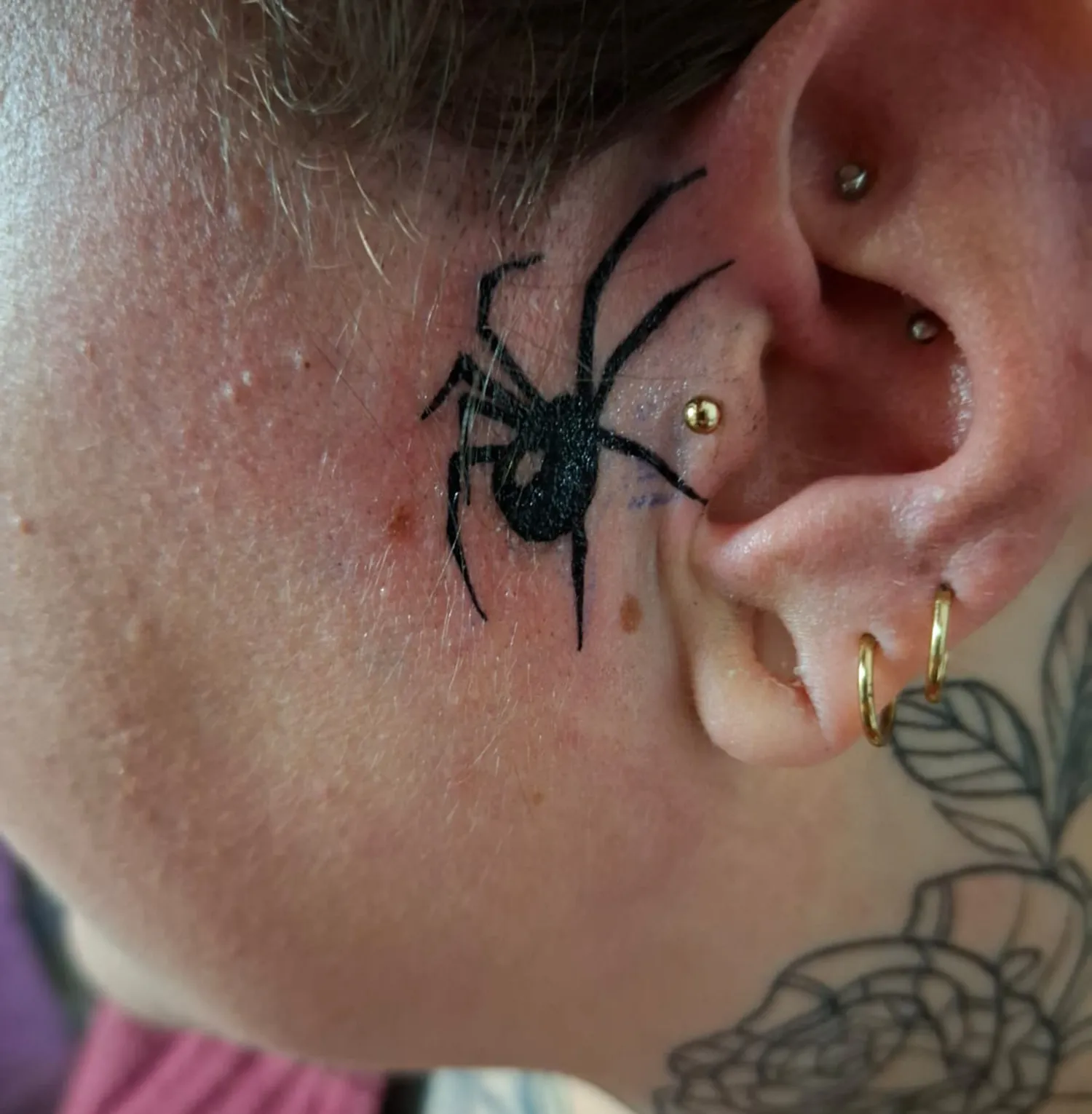 Feines Spinnentattoo hinter dem Ohr mit goldenen Piercings und floralen Linien im Hintergrund
