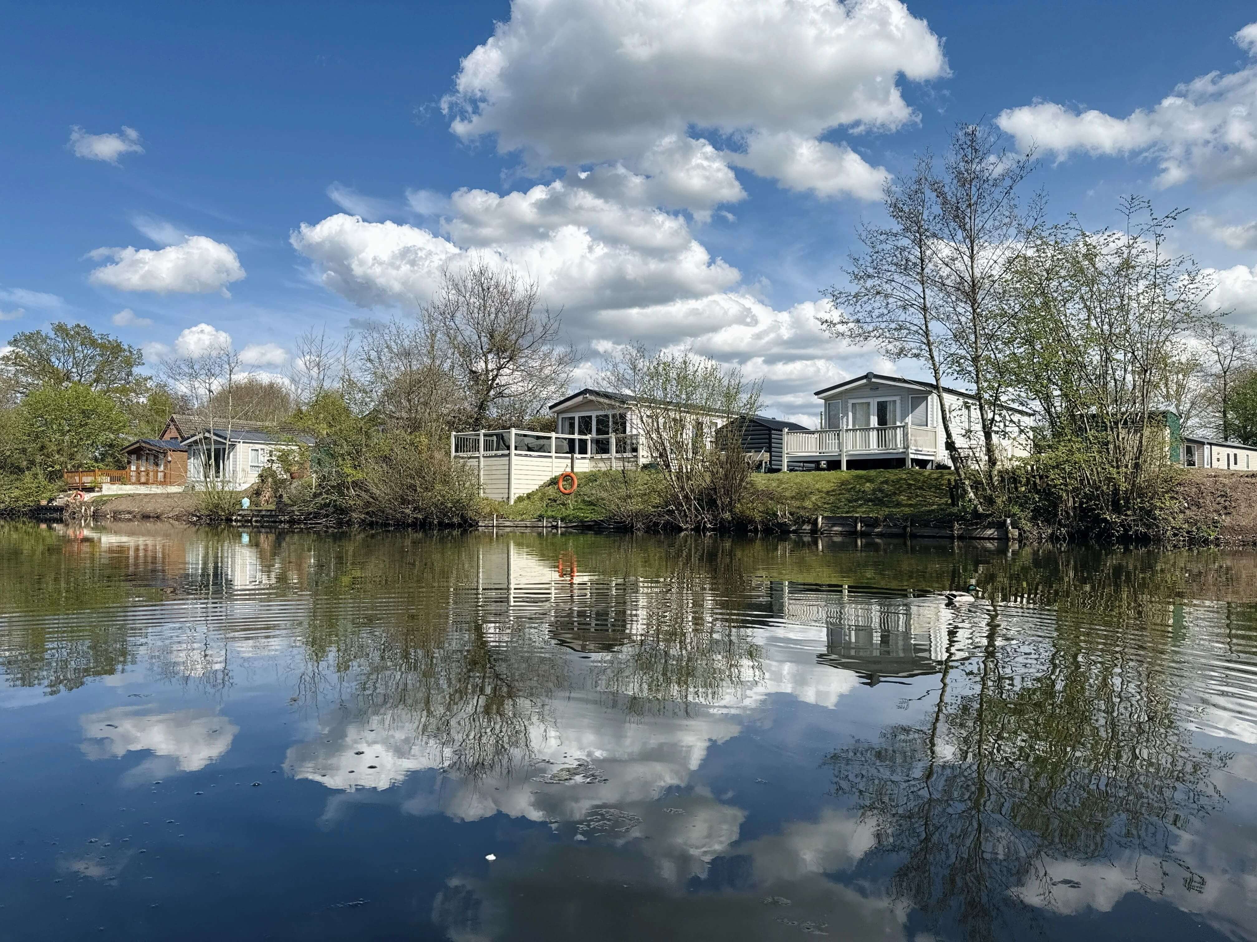 Lakeside holiday park with white cabins reflecting in calm water under cloudy sky