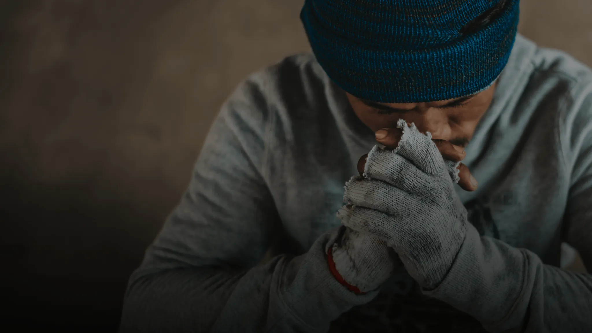 Man sitting with clasped hands and lowered head, wearing a hat and gloves