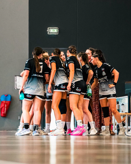 Plusieurs joueuses du Pouzauges Vendée Handball, en tenue noire et blanche, se regroupent sur le terrain avant ou pendant un match de handball. 