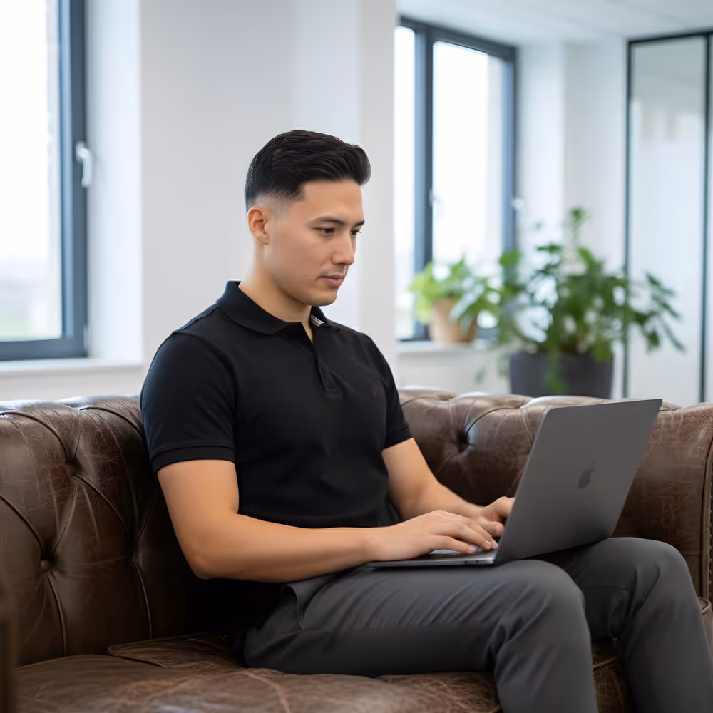 Man zittend op een bruine leren bank, werkend op een laptop in een lichte kamer met ramen en groene planten.
