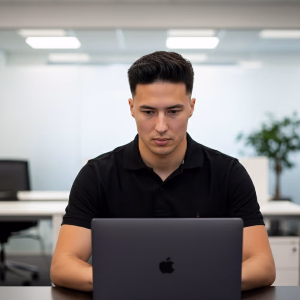 Man in zwart poloshirt werkt geconcentreerd op een MacBook in een moderne kantooromgeving.
