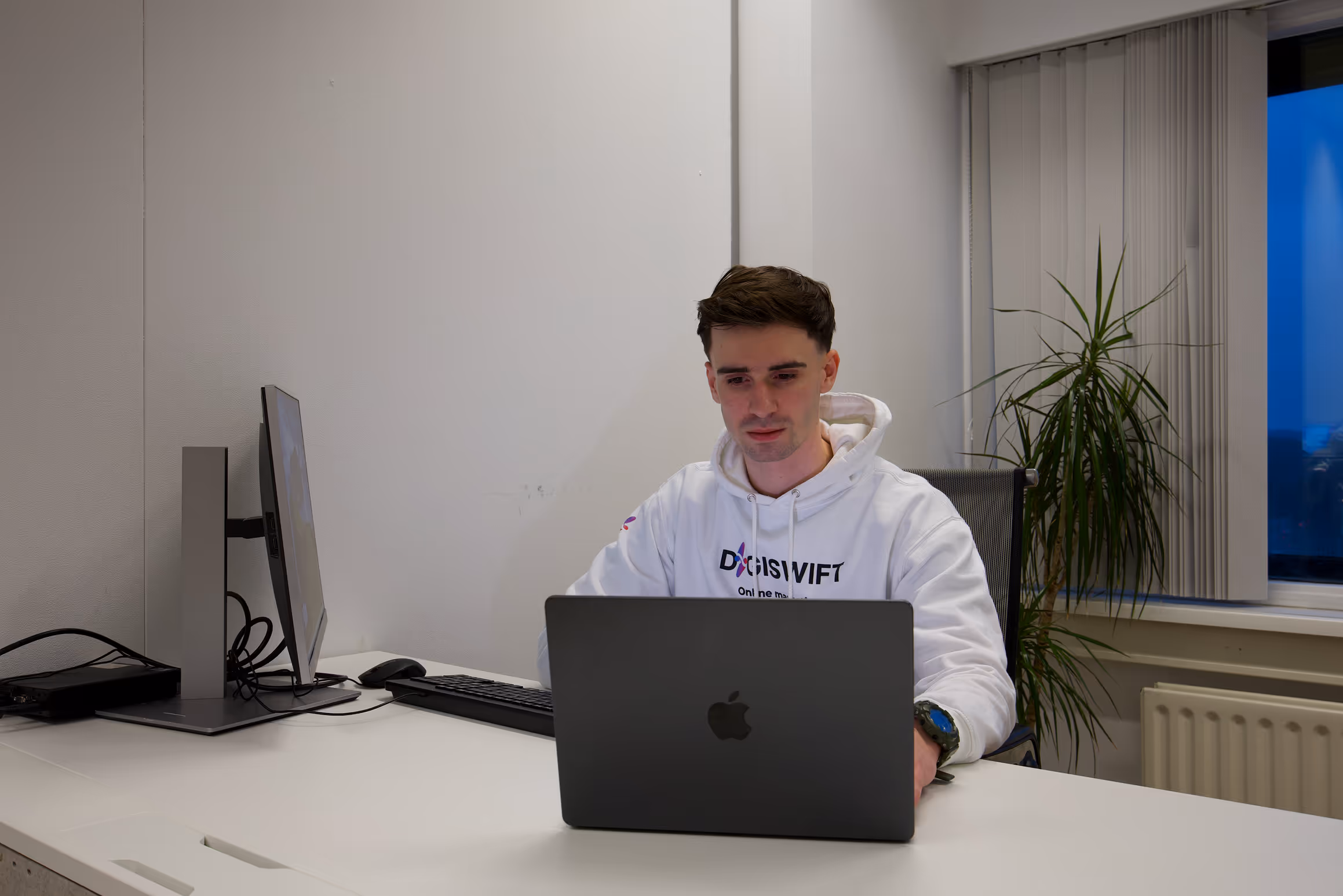 Daniel Coenen, mede-eigenaar en Webflow developer bij DigiSwift, zit achter bureau met laptop en extern beeldscherm in kantoorruimte met raam en plant op de achtergrond