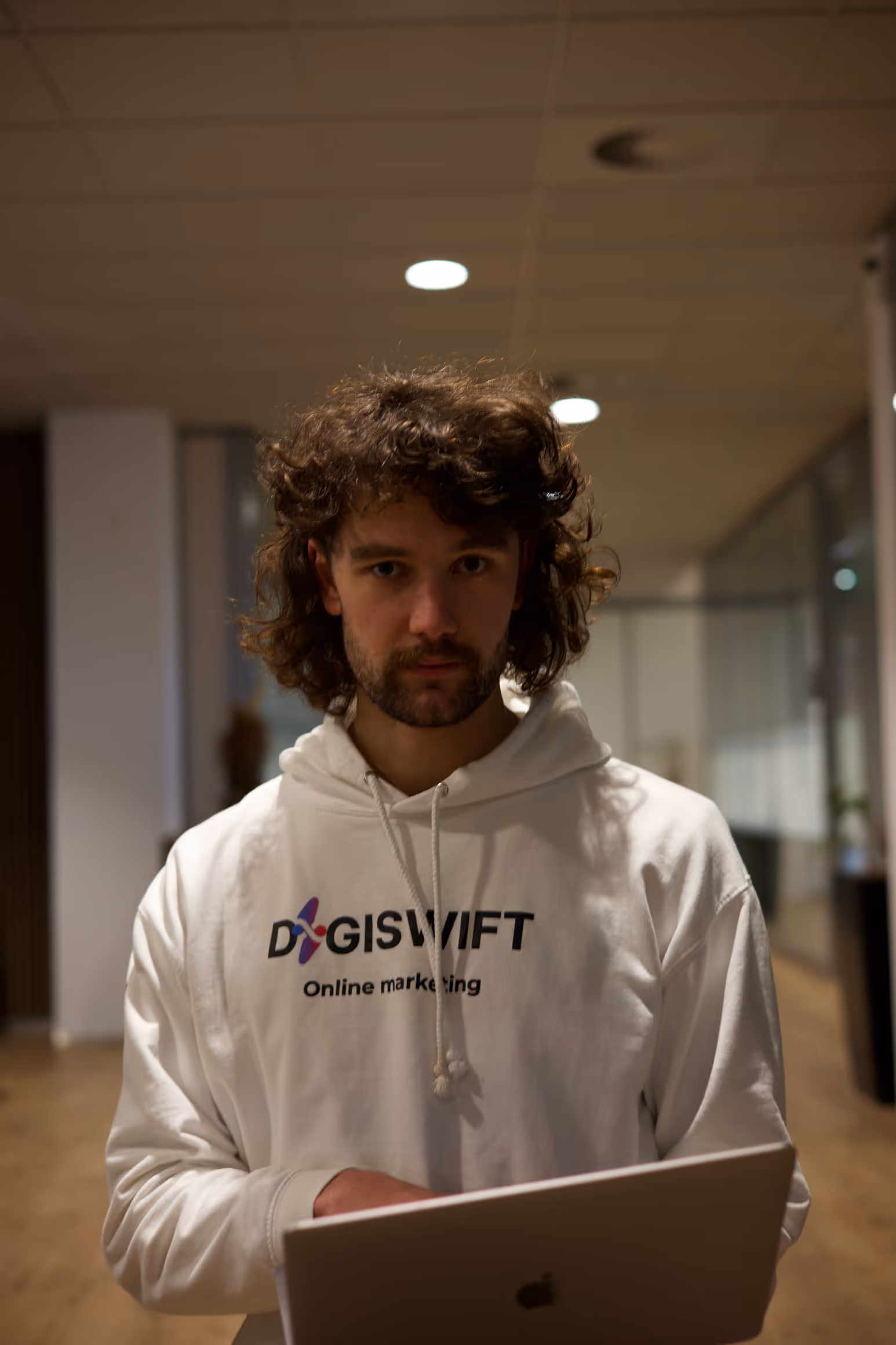 Jon Rijnsburger, webdesigner bij DigiSwift, staat in witte DigiSwift hoodie met laptop in handen in kantooromgeving en kijkt recht in de camera