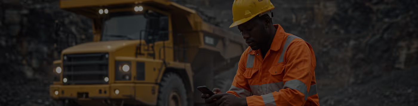 A Black mine worker wearing a yellow hard hat and orange high-visibility overalls looks at their phone, with a large yellow mining truck in the background.