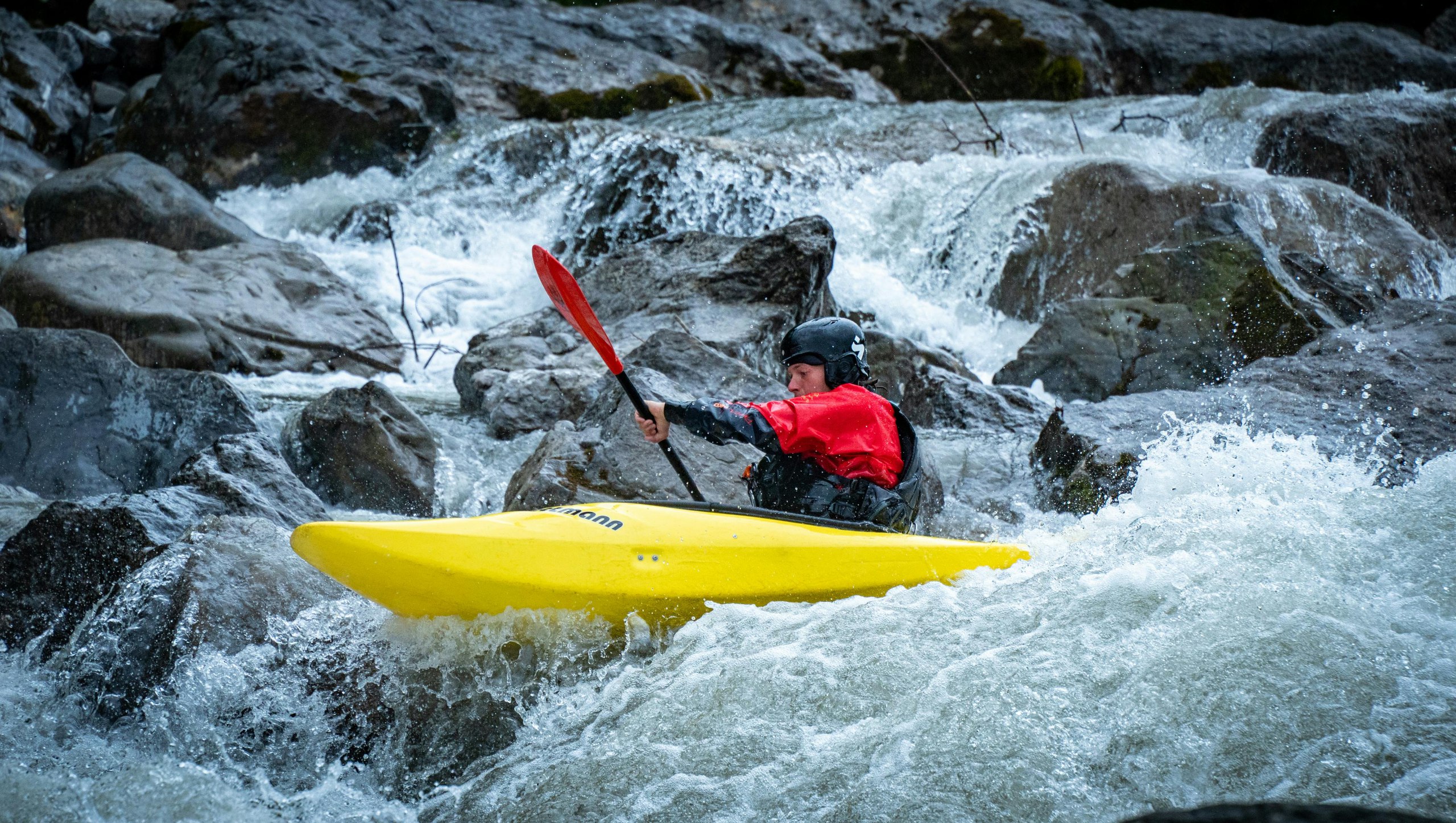 Person in a yellow kayak wearing a red jacket and black helmet paddling through whitewater rapids surrounded by large rocks