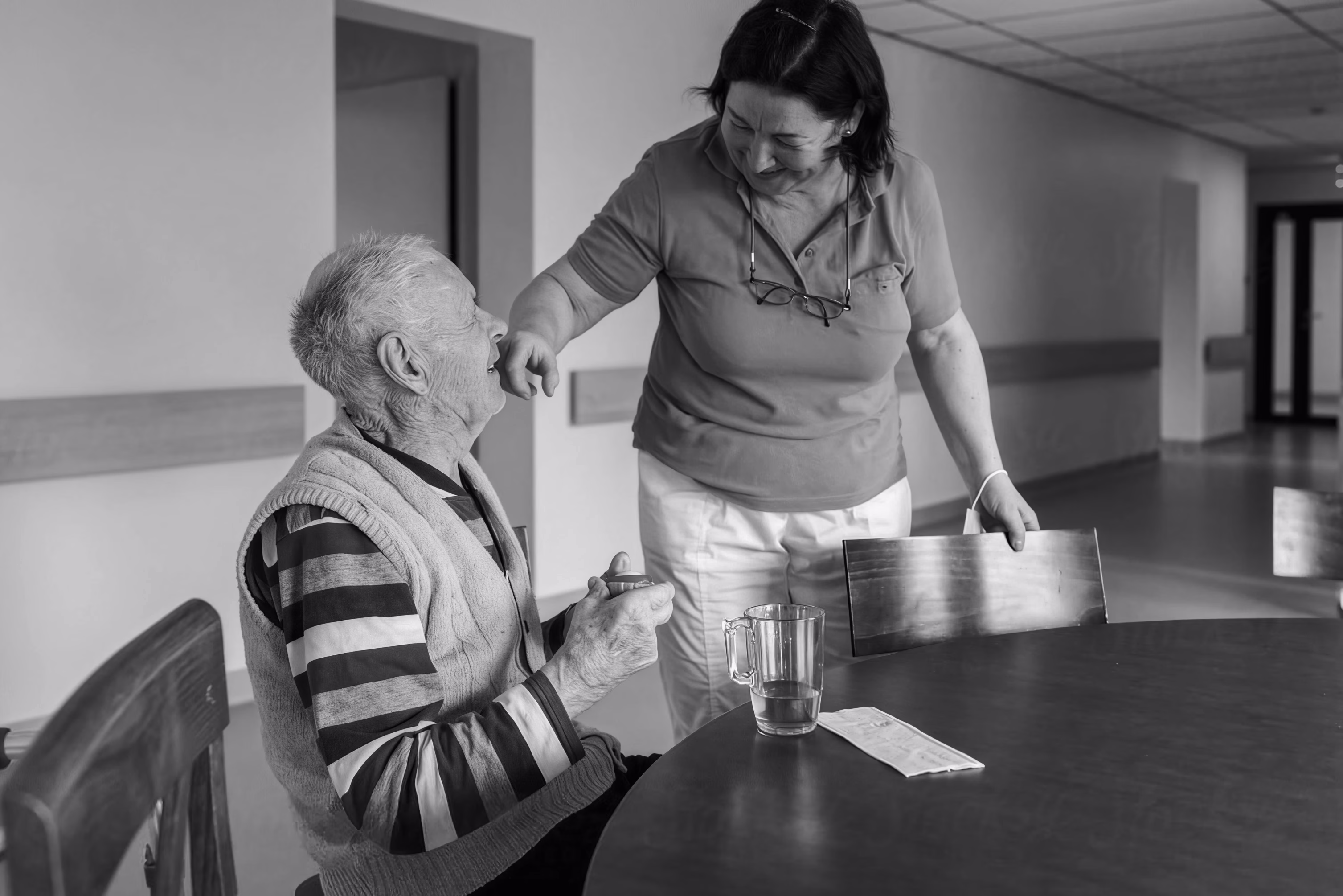 Caregiver serving a drink to an older man seated at a table in a skilled nursing facility.