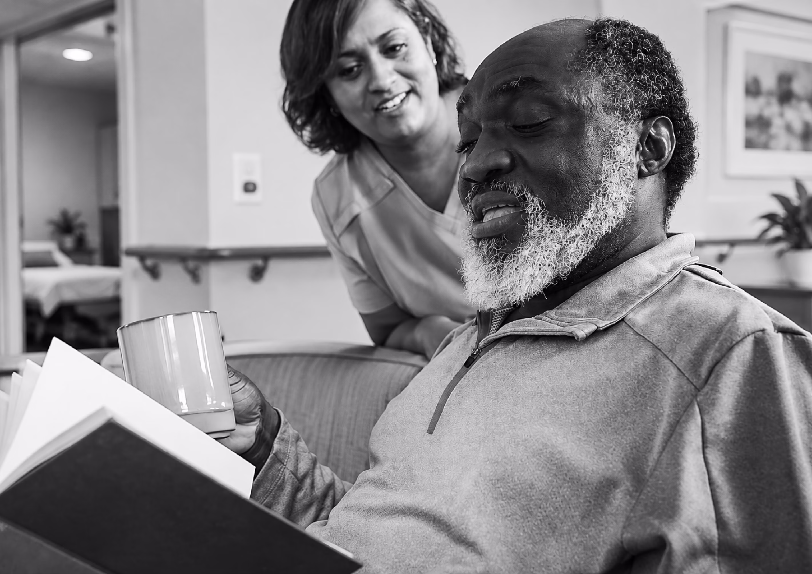 Caregiver talking with an older man as he reads in a care facility.