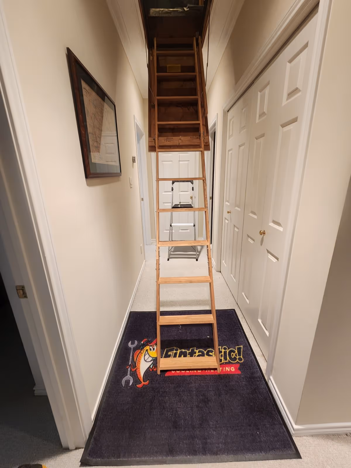 Wooden ladder leading to attic in narrow white hallway with framed art