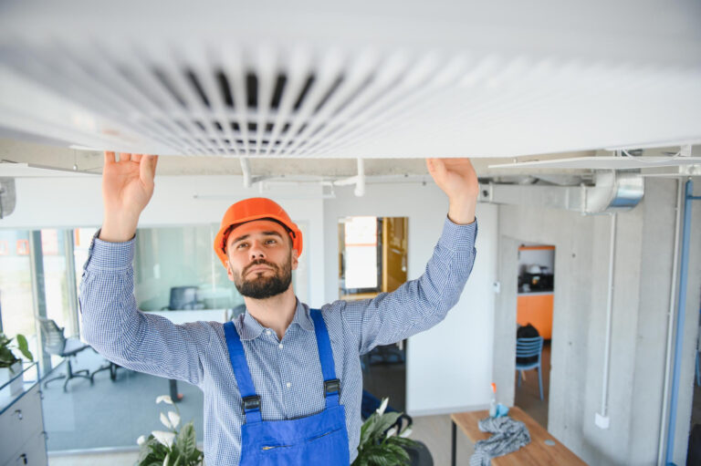 A bearded male HVAC technician wearing an orange hard hat and blue overalls is reaching up to inspect a ceiling-mounted commercial air conditioning vent.