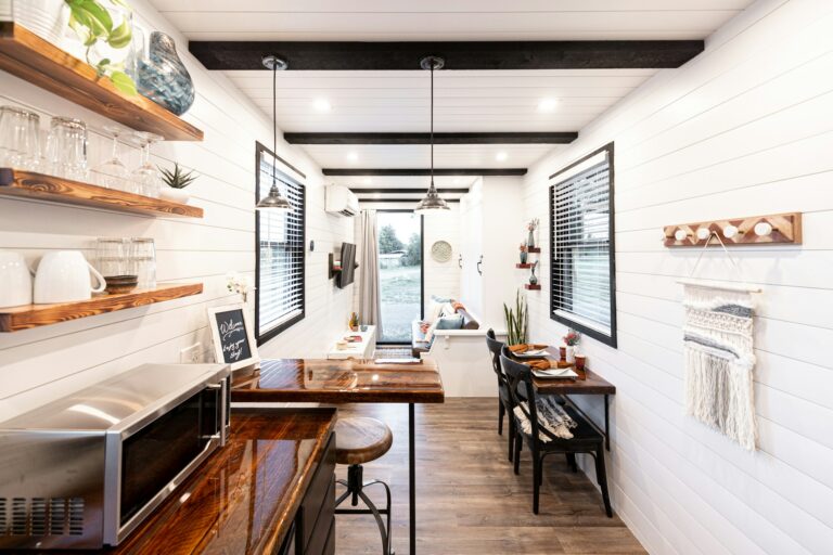 A modern, minimalist interior of a tiny home with a kitchen, living area, and dining space, featuring white walls and wooden accents.