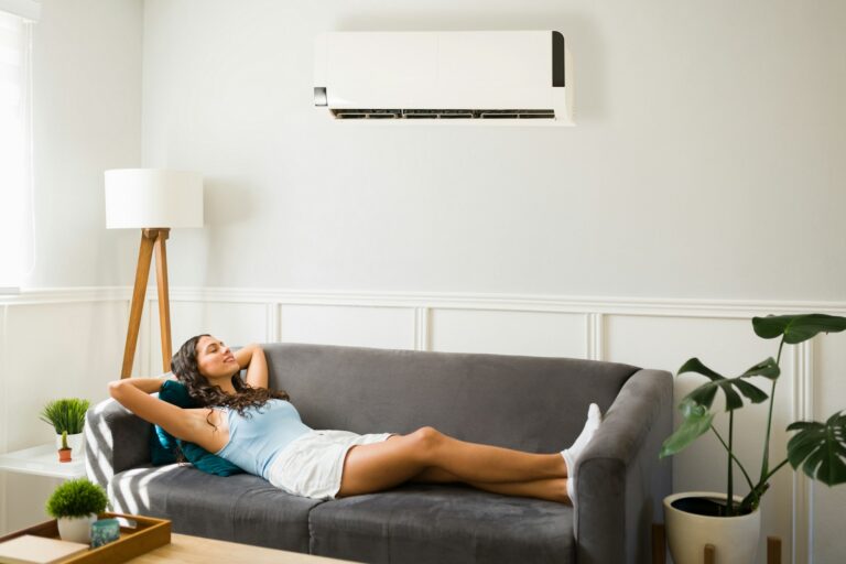  A woman is relaxing on a gray sofa in a living room, with a ductless mini-split air conditioner mounted on the wall above her.