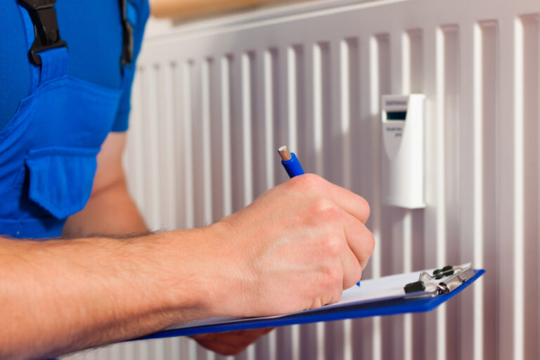 A male heating engineer wearing blue overalls and holding a clipboard inspects a white radiator with a thermostatic valve.