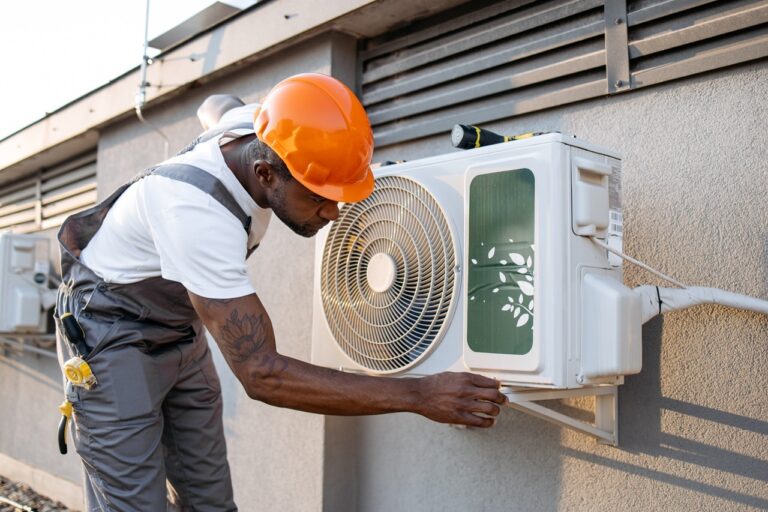  A technician wearing an orange hard hat and gray overalls is inspecting an outdoor air conditioning unit mounted on the side of a building.