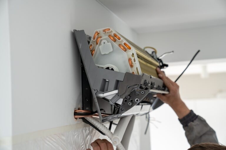 A professional is working on the internal components of a wall-mounted ductless air conditioner, which is partially disassembled to show its copper coils and fan.