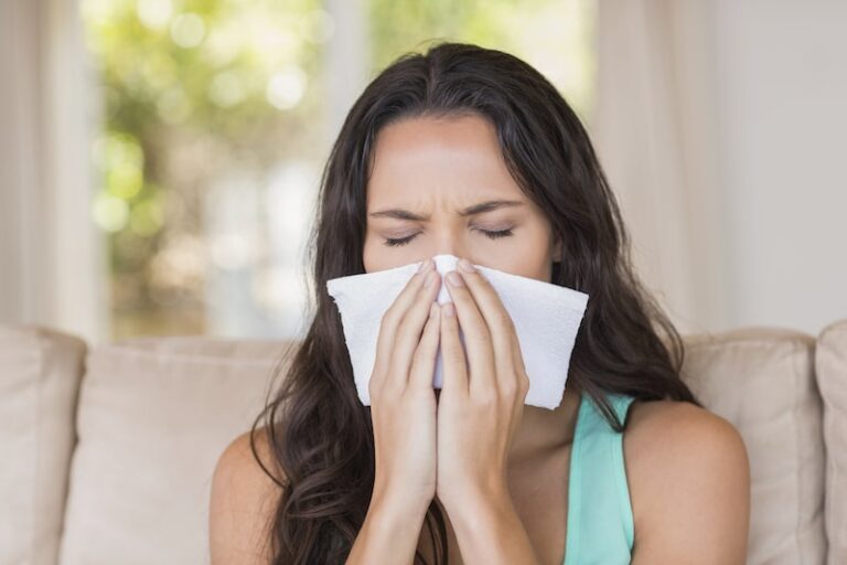  A woman with long, dark hair is blowing her nose with a tissue, with her eyes closed in discomfort.