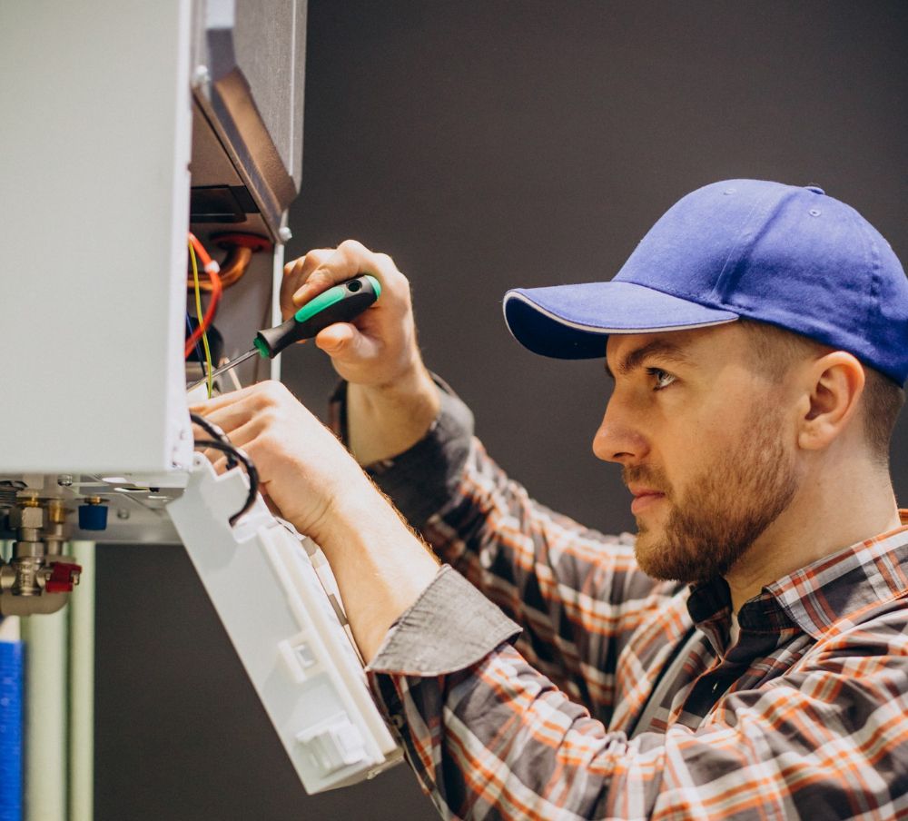 A focused technician wearing a blue cap and a plaid shirt uses a screwdriver to work on the internal wiring of a gas boiler.