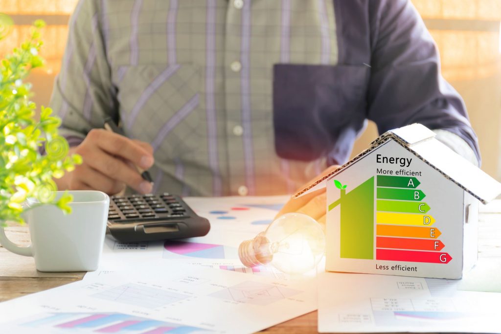 A person sits at a desk with a calculator and papers, next to a small cardboard house with an energy efficiency rating chart from A to G.