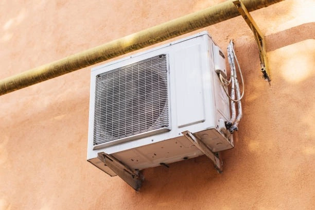 An outdoor mini-split air conditioning unit is mounted on a pink stucco wall below a yellow pipe.