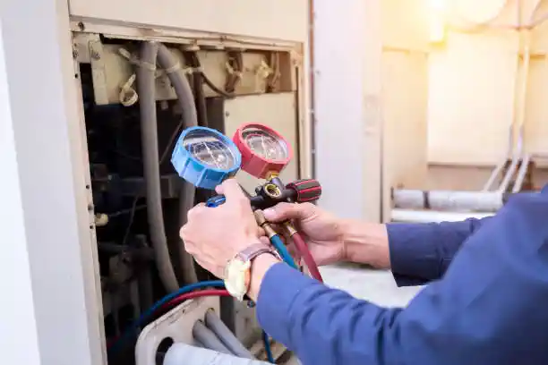  A man in a blue shirt works on an HVAC unit, using a manifold gauge set to check the refrigerant levels. His hands are visible holding the tool with blue and red dials and hoses connected to the system.