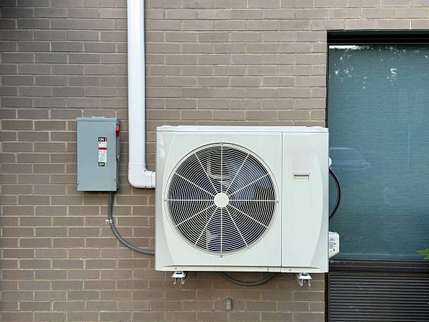 A light-gray mini-split air conditioning condenser unit, with a circular fan grill, is mounted on a brown brick wall. A gray electrical box with "ON" and "OFF" labels is attached to the wall to the left of the unit,
