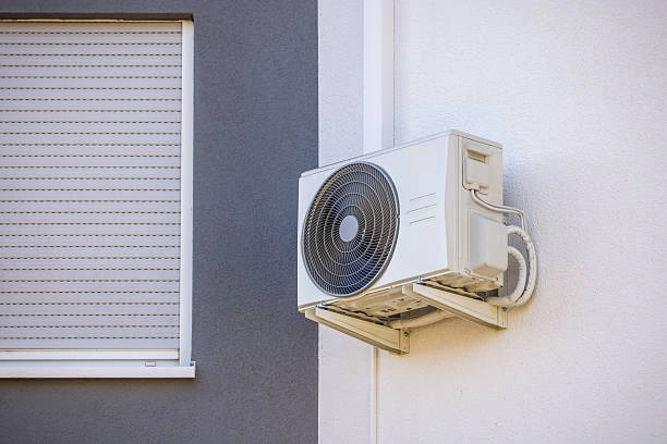  A light-colored mini-split air conditioner condenser unit is mounted on a white wall. The unit has a circular fan grille and is positioned to the right of a window with closed horizontal blinds.