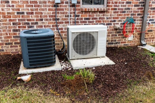  A traditional cylindrical air conditioning unit and a rectangular mini-split outdoor unit sit on concrete pads next to a brick building. The ground around the units is covered in dark mulch, with a patch of grass in the foreground. 