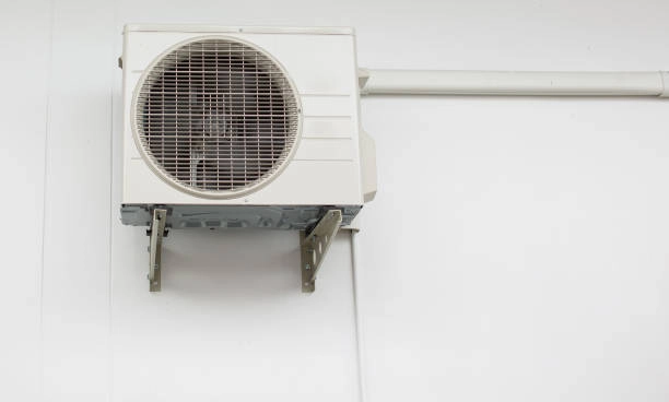 A small, dirty air conditioning unit is mounted on a white wall. The unit's fan and grille are visible, and it is supported by two metal brackets. A white pipe runs horizontally along the wall from the right side of the unit.