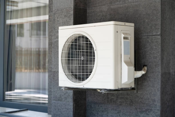 A light-colored, mini-split air conditioner condenser unit is mounted on a dark gray tiled wall. The unit has a circular fan grille on the front and is attached to the wall by brackets. A window with a gray frame is visible to the left.