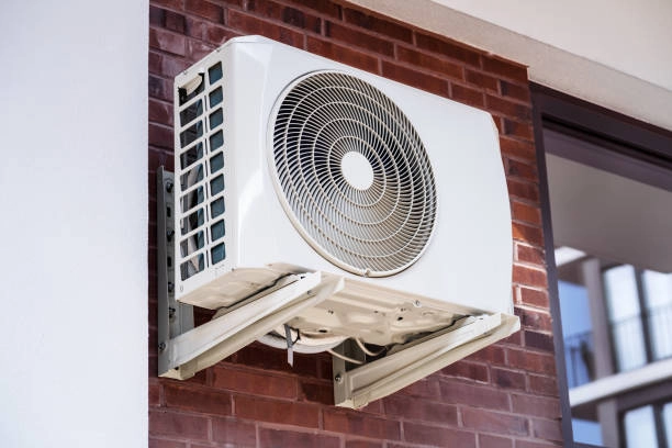 A wall-mounted white air conditioner condenser unit, with a circular fan grill, is attached to a red brick wall. The front panel is unlatched and tilted downward, exposing the internal components.