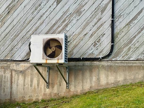 A weathered white mini-split air conditioner condenser unit is mounted on a concrete foundation below a wall of peeling gray-and-white diagonal siding.