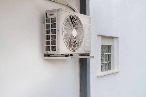 A mini-split air conditioner condenser unit is mounted on a white wall. The unit's cover is open, exposing the inside of the unit and the circular fan grille.
