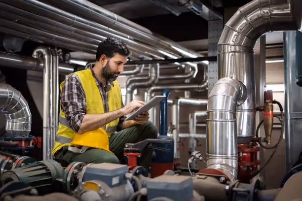 Worker in safety vest checks industrial equipment using tablet among pipes