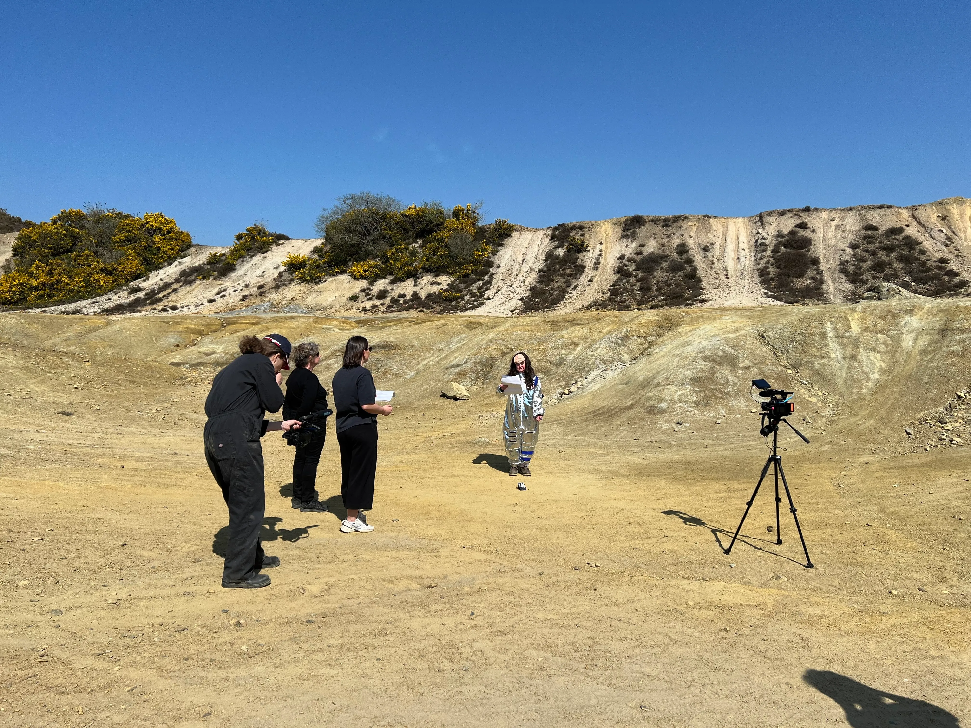 a photo of a film shoot where an actor is wearing a silver spacesuit in a sandy quarry with people standing around filming