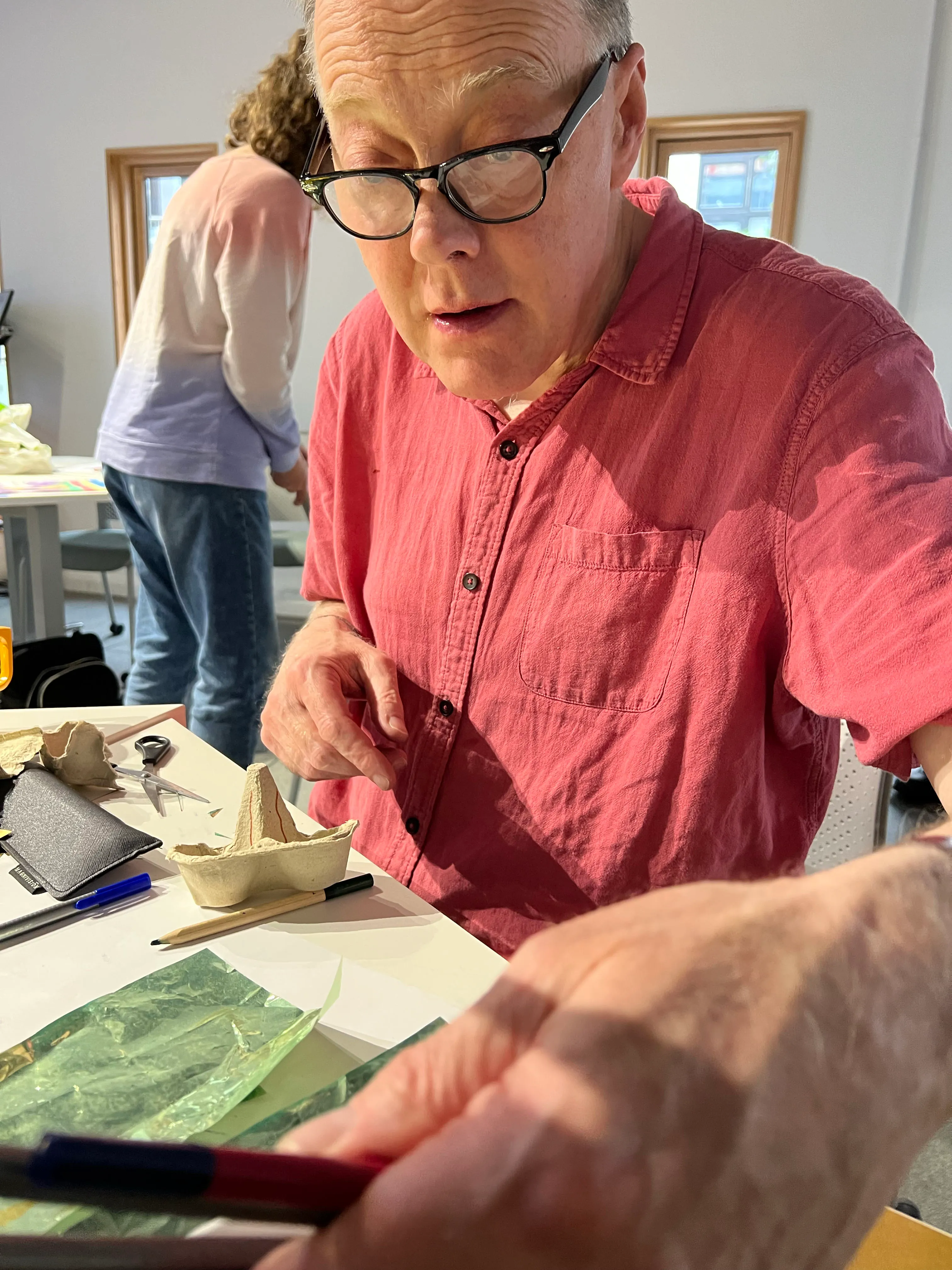 A photo of a man in a red shirt drawing at a workshop table and reaching for a new pen