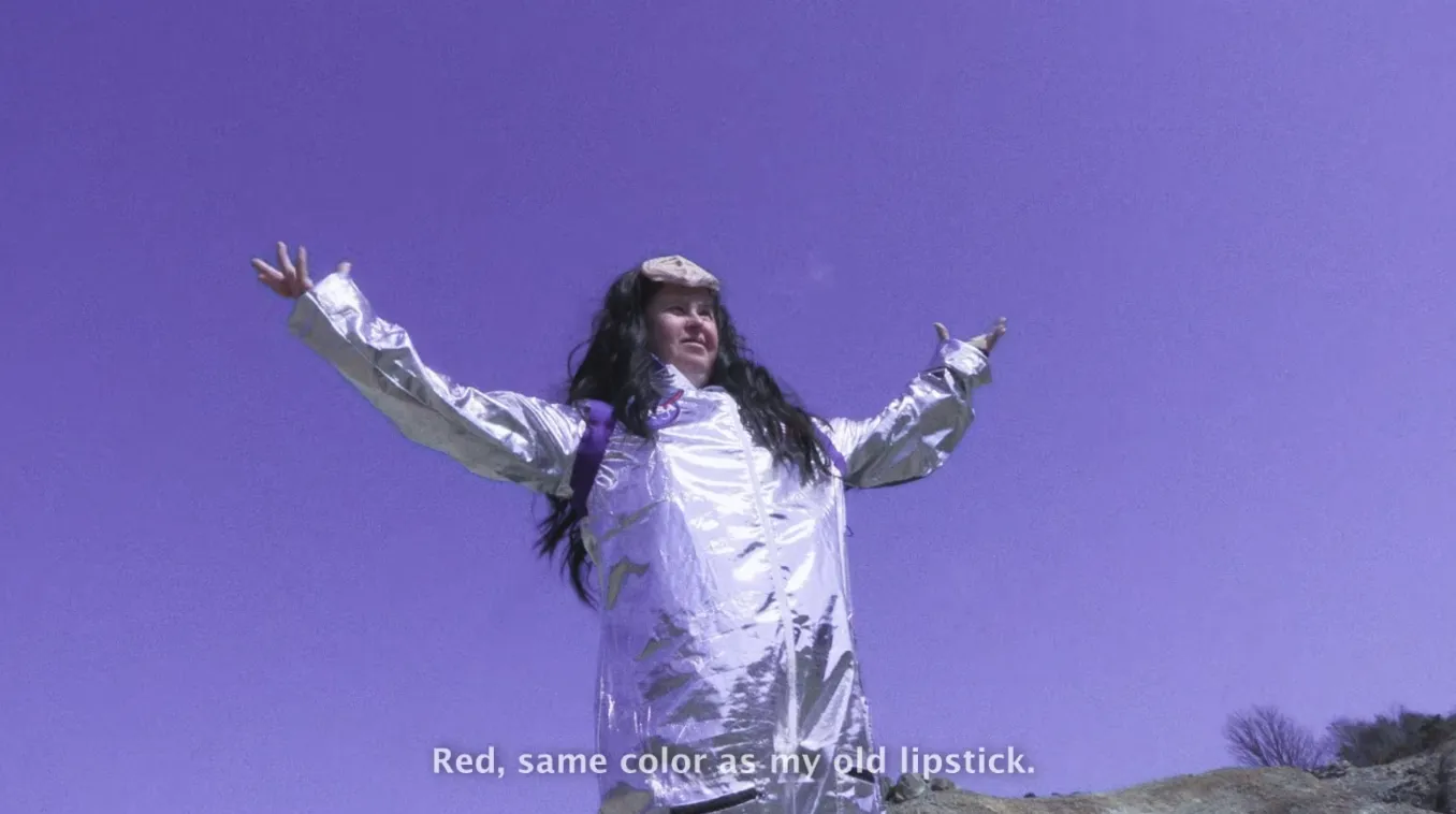 A still from a film with a person wearing a silver spacesuit and a long brown wig and raising their arms wide like a bird under a deep blue sky.