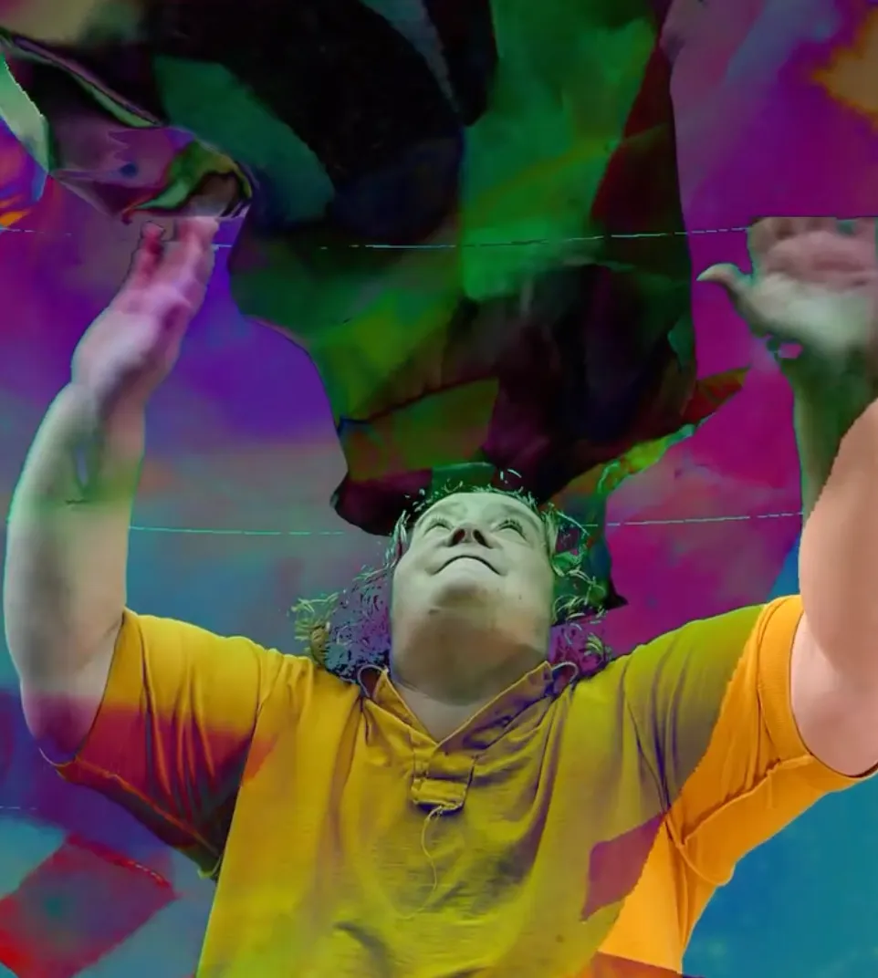 a still from a film with a woman wearing a yellow teeshirt with her hands raised above her head. She is standing in front a multi colour background 