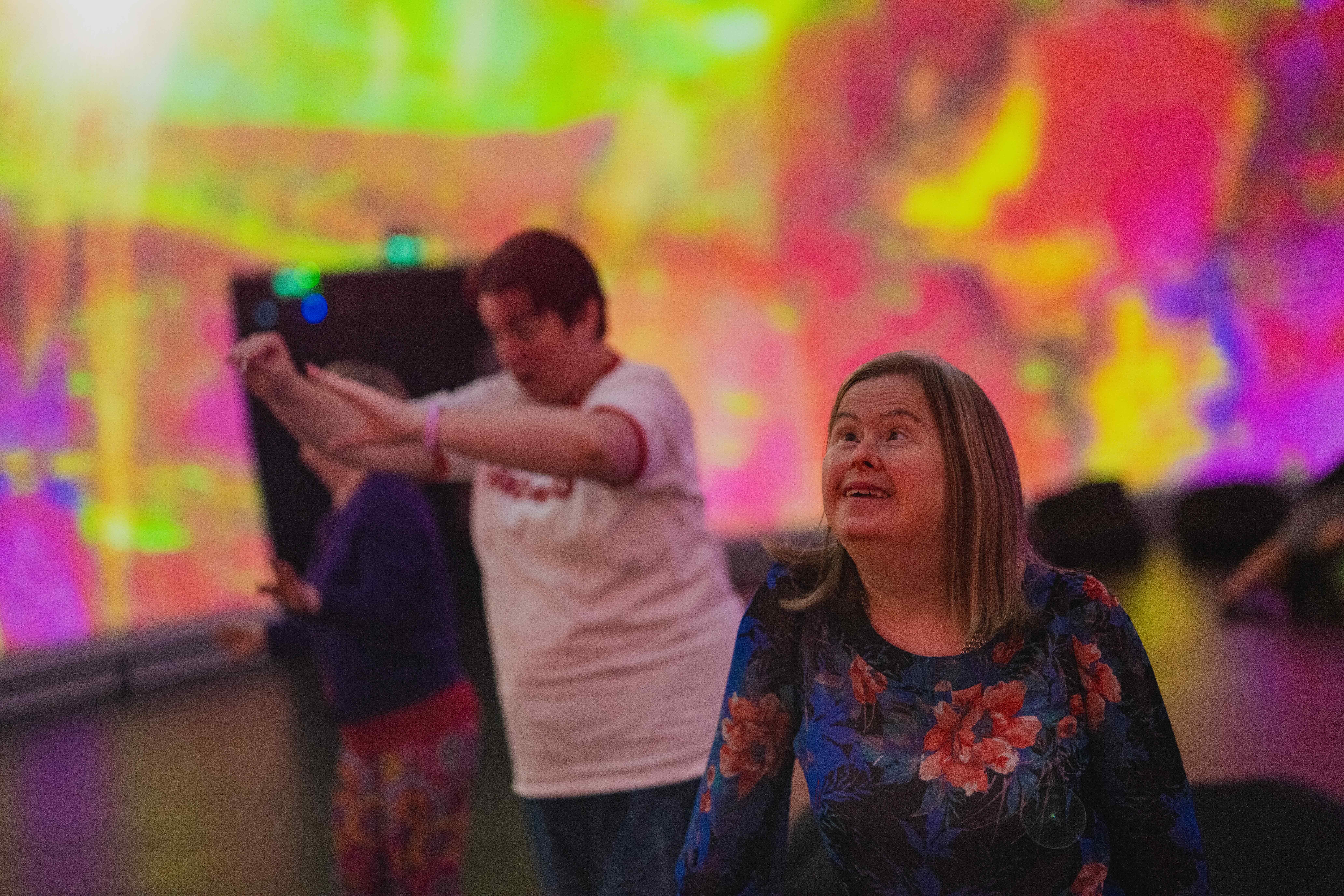 A photo of a woman wearing glasses raising her arms out and looking up at a purple and pink film projection all around her on the wall