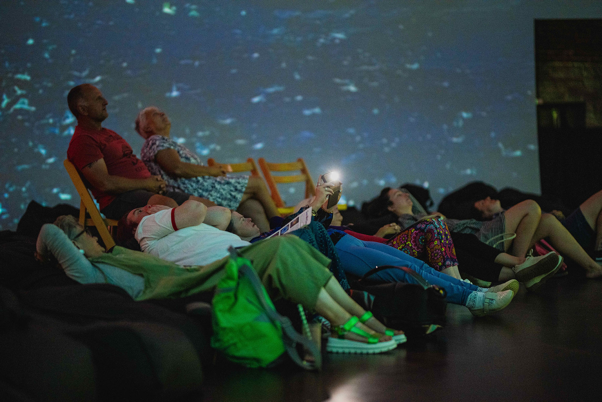 a dark 360 cinema with a light blue and white spot projection on the wall behind an audience of people looking up sitting on bean bags and 2 chairs