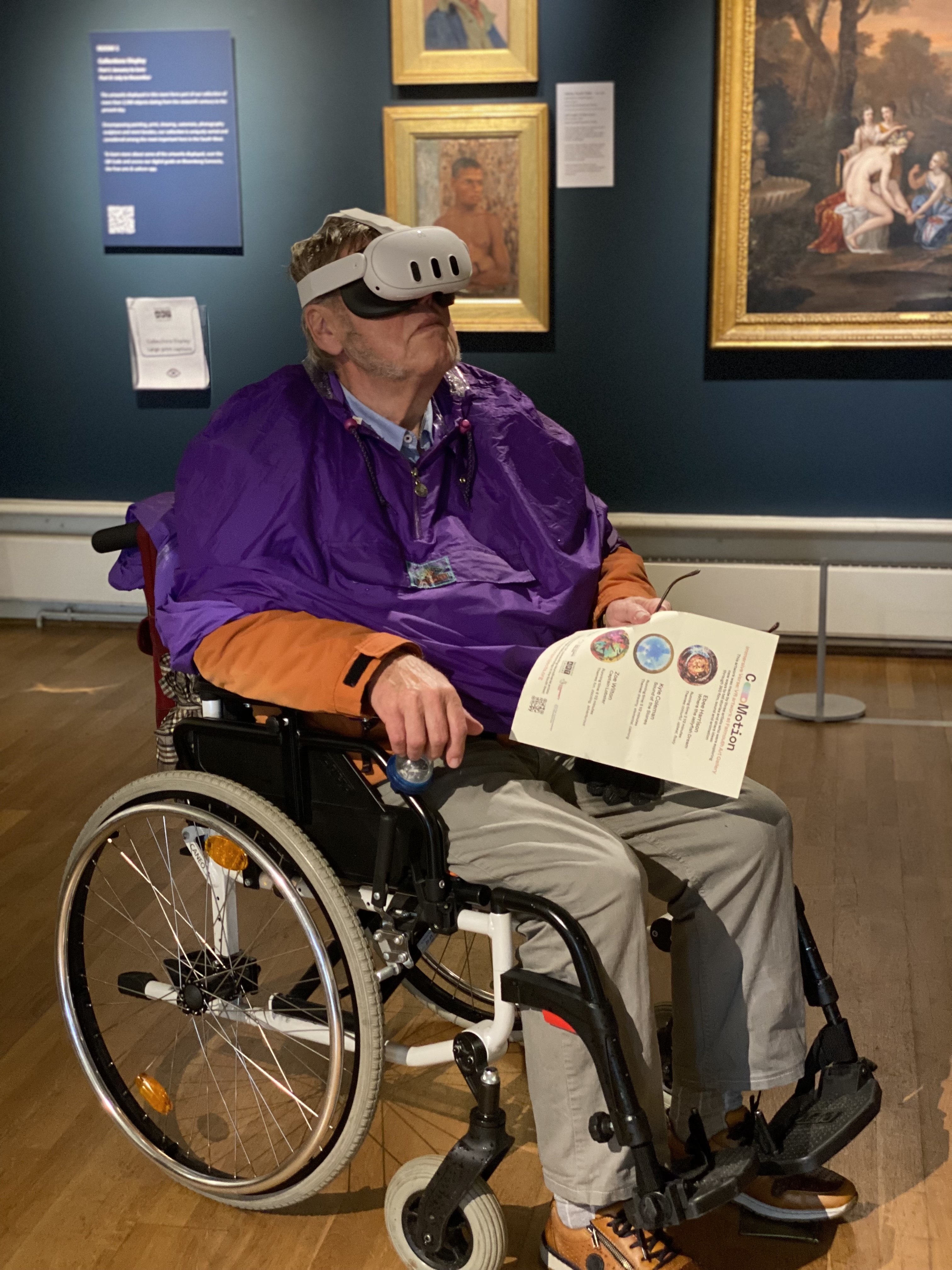 A photo of a white man in his 70's, wearing a purple top and greay trousers, he is in a wheelchair and wearing a VR headset. The background is in a museum with gold framed paintings on a dark blue wall.