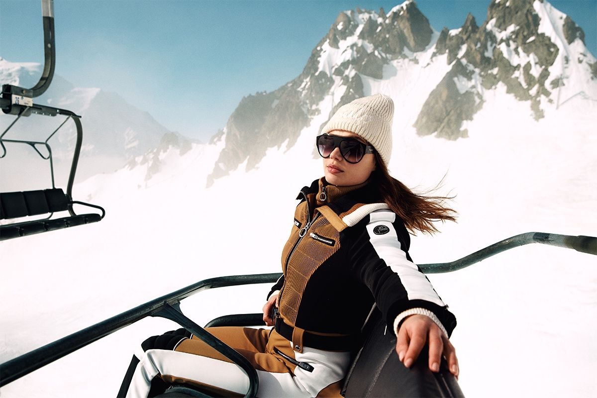 Femme portant une tenue de ski Fusalp sur un télésiège, avec un paysage de montagnes enneigées en arrière-plan.