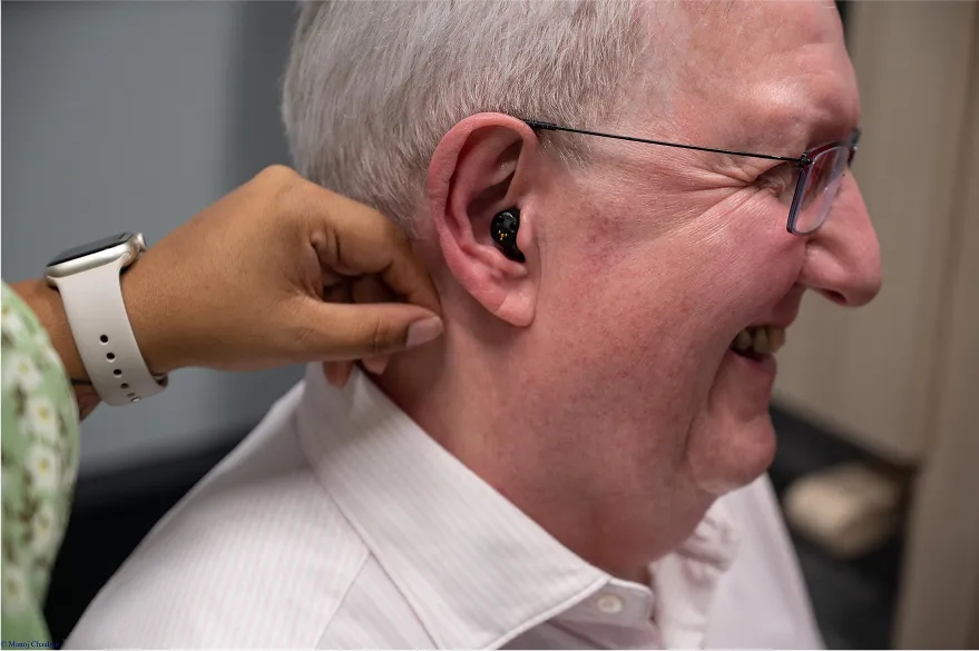 Hearing specialist fitting hearing aid for smiling older man with glasses.