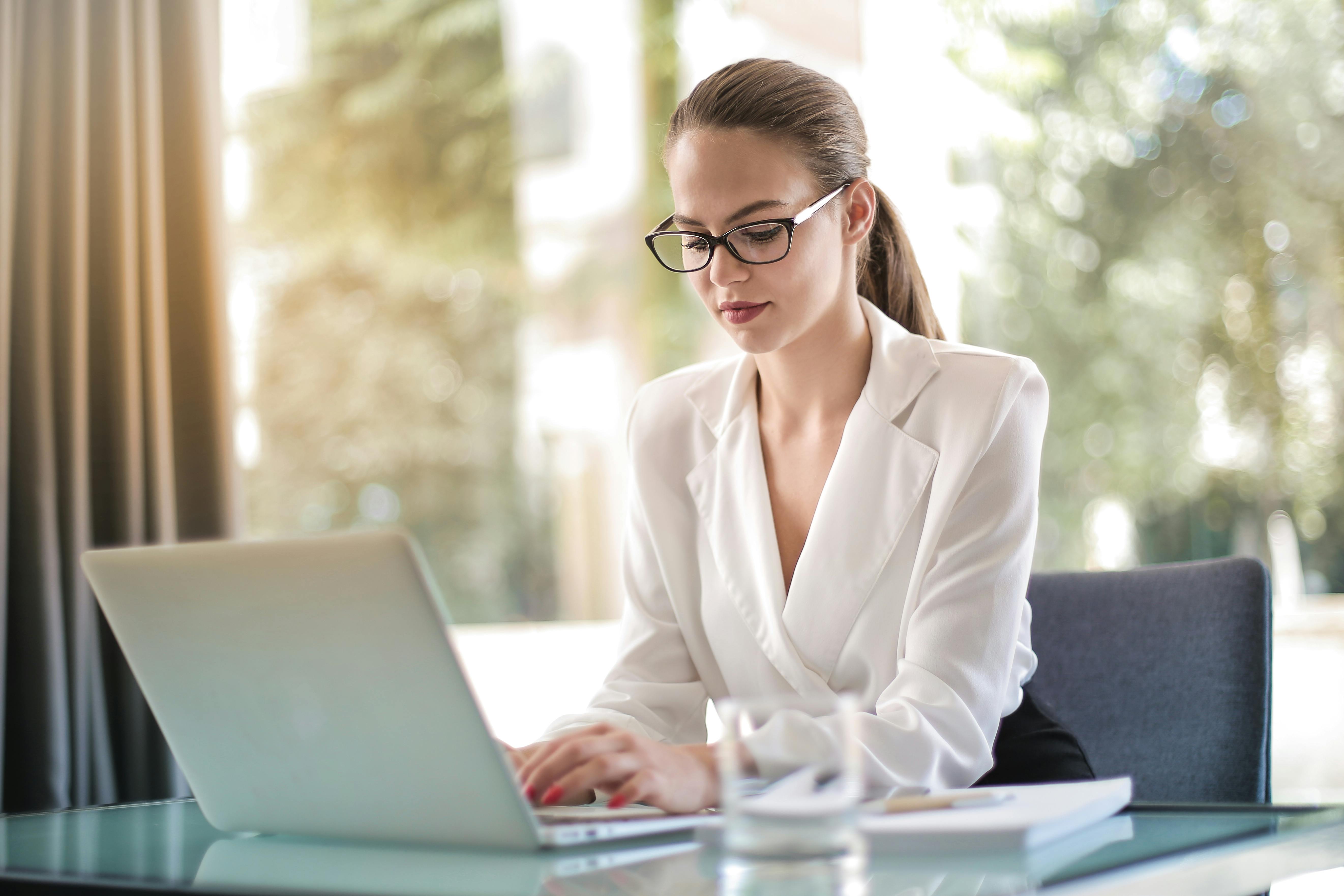 Frau im weißen Blazer und mit Brille arbeitet an einem Laptop an einem Glas-Schreibtisch.