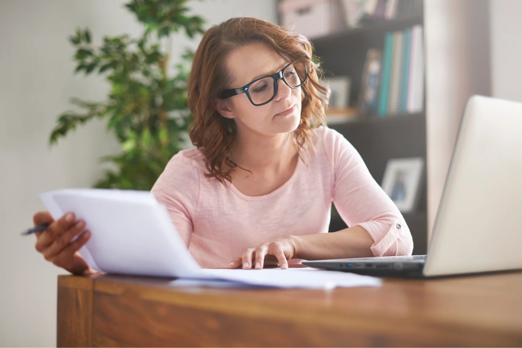 Frau mit Brille arbeitet konzentriert mit Dokumenten und Laptop am Holztisch.