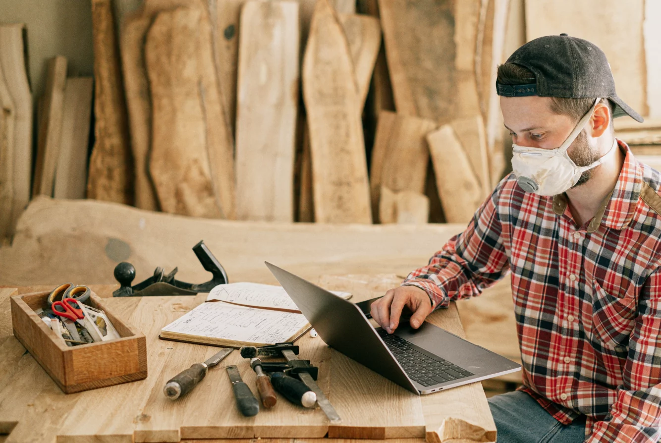 Mann mit Mundschutz und kariertem Hemd arbeitet an einem Laptop in einer Holzwerkstatt mit Werkzeugen auf dem Tisch.