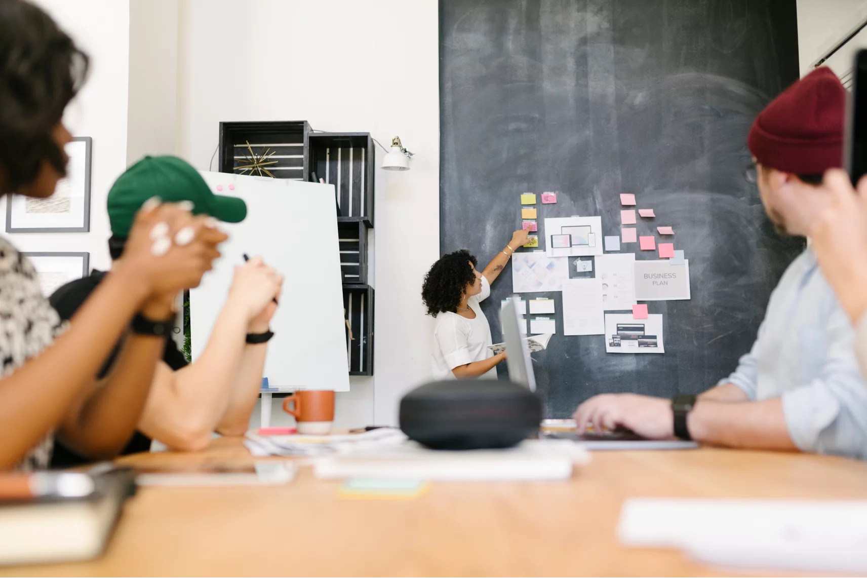 Person präsentiert Ideen an einer Tafel mit Notizzetteln, während eine Gruppe von vier Personen am Tisch aufmerksam zuhört.