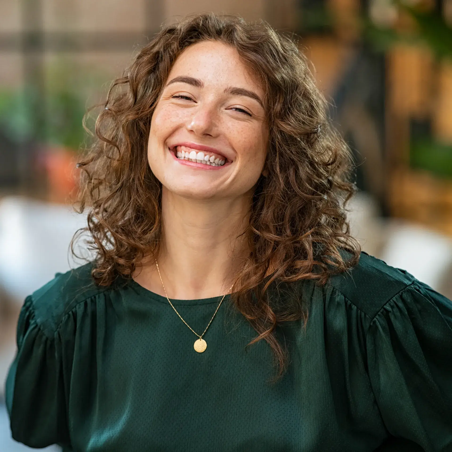 A woman with curly hair smiles brightly while wearing a dark green top and gold necklace.