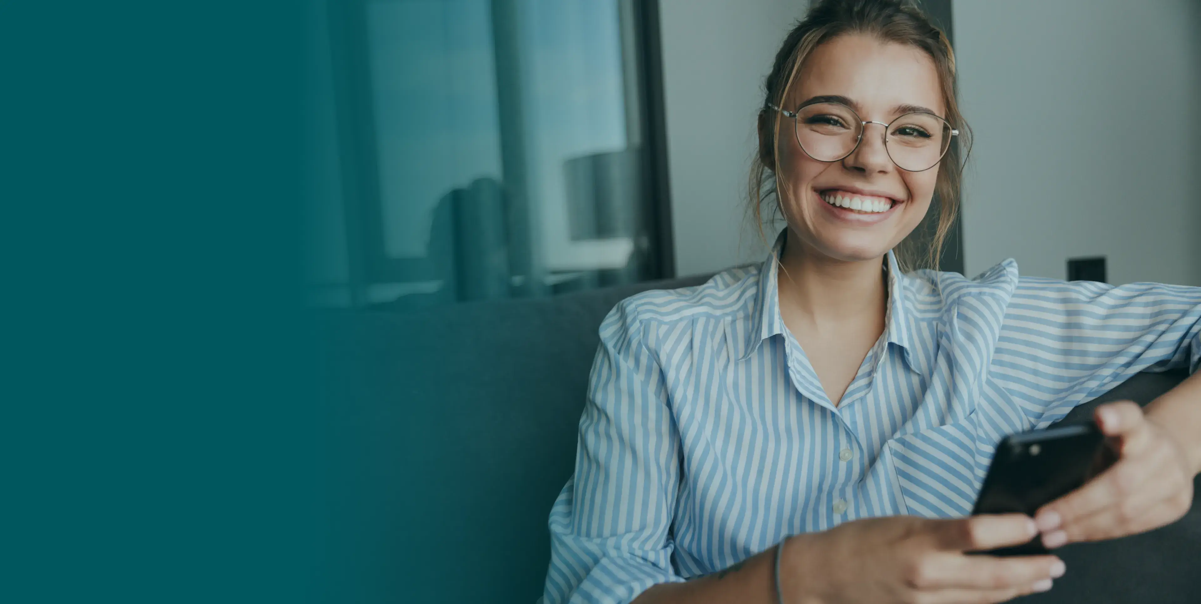 Woman wearing glasses, smiling broadly while sitting on a couch and holding a smartphone.