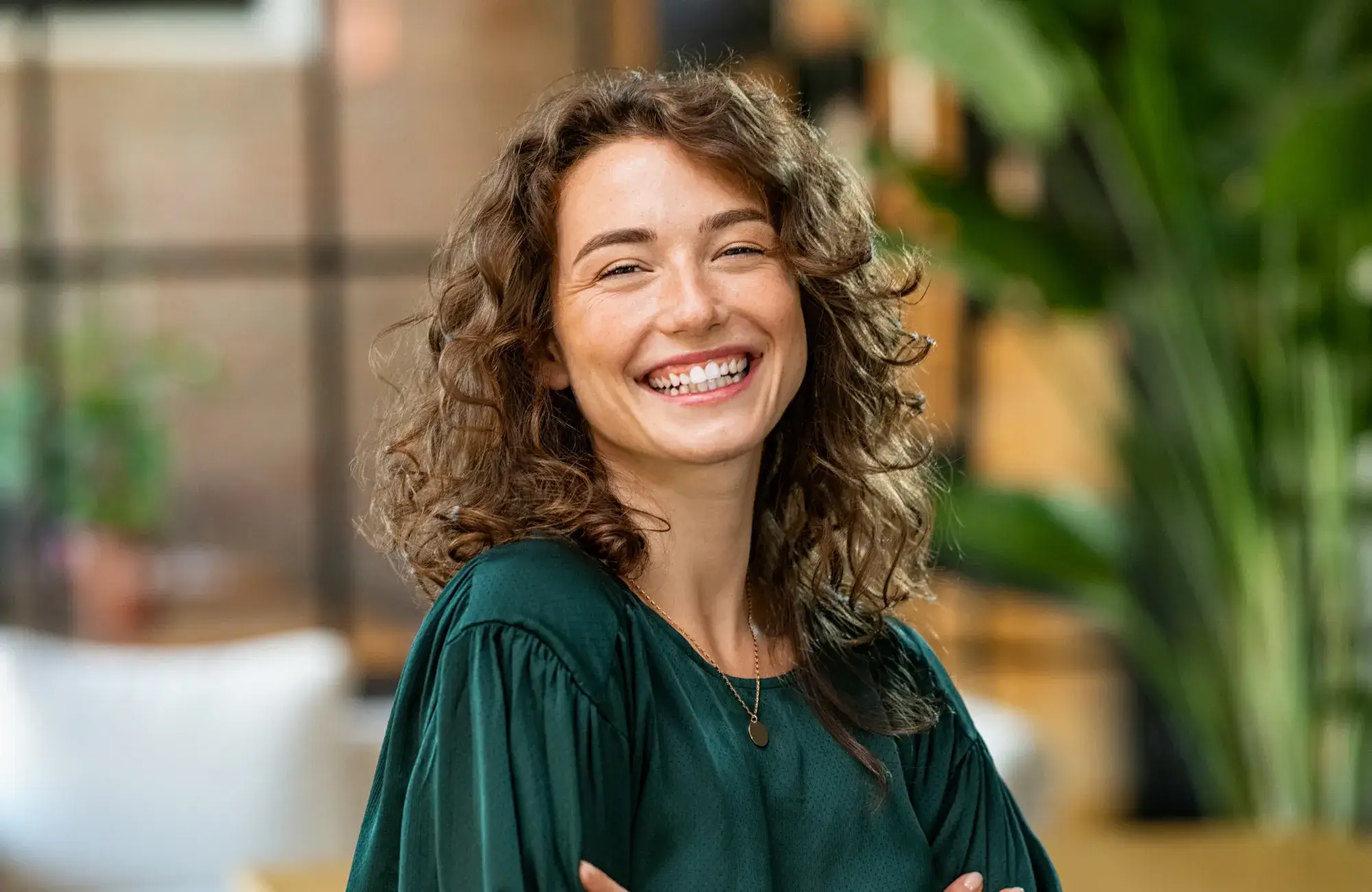 Smiling woman with curly hair wearing a green top stands indoors.